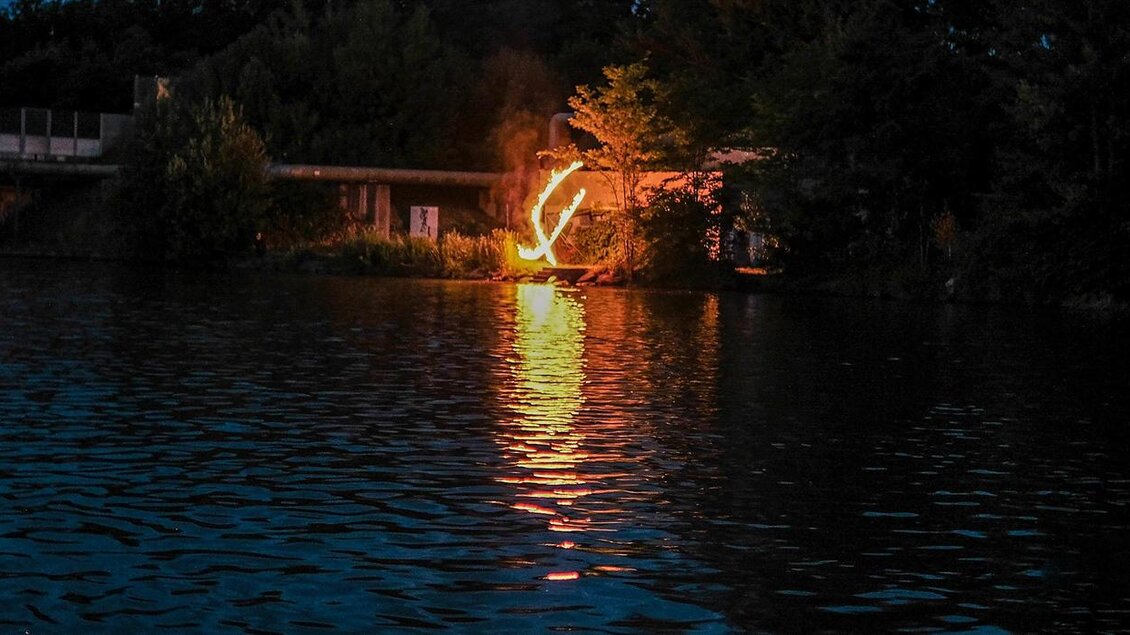 Ein ruhiger See bei Nacht mit spiegelndem Wasser. Im Hintergrund leuchtet ein Feuer und erhellt die Umgebung. | © Austria eXtreme