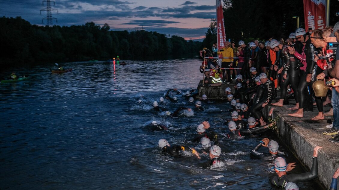 Eine Gruppe von Schwimmern steht am Ufer und bereitet sich auf ein Wettkampf im Wasser vor. Im Hintergrund ist ein Fluss und ein dunkler Himmel zu sehen. | © Austria eXtreme