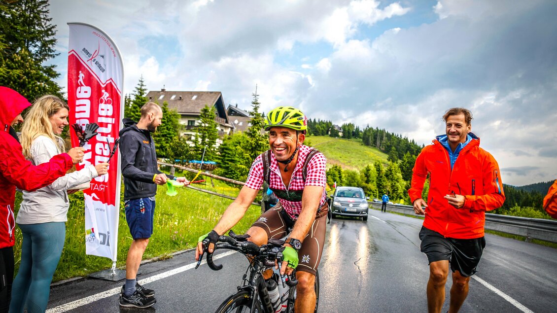 Ein Radfahrer lächelt beim Rennen auf einer nassen Straße. Im Hintergrund stehen Zuschauer und es gibt eine schöne grüne Landschaft. | © Austria eXtreme