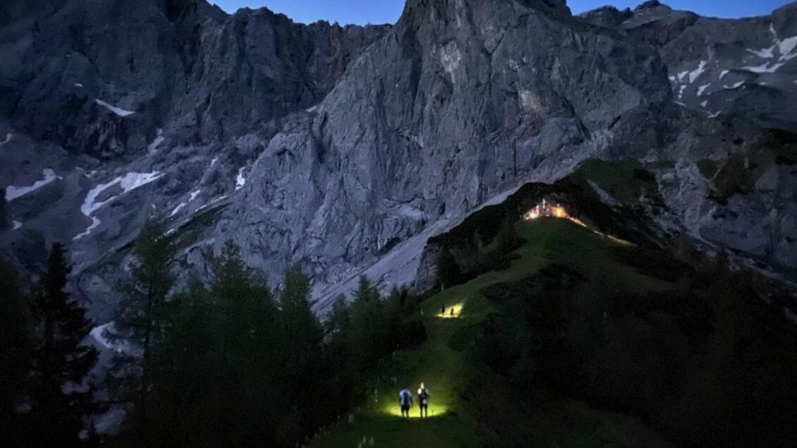 Zwei Wanderer mit Taschenlampen erklimmen einen Bergpfad in der Dämmerung. Majestätische Felsen und Bäume umgeben die Szene. | © Austria eXtreme