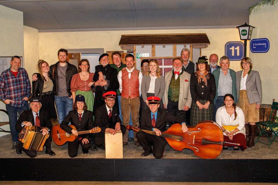 Eine Gruppe von Menschen posiert auf einer Bühnenkulisse. Einige tragen traditionelle Kleidung und halten Musikinstrumente. | © Erich Kainzinger