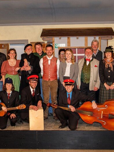 A group of people poses against a stage backdrop. Some are wearing traditional clothing and holding musical instruments. | © Erich Kainzinger