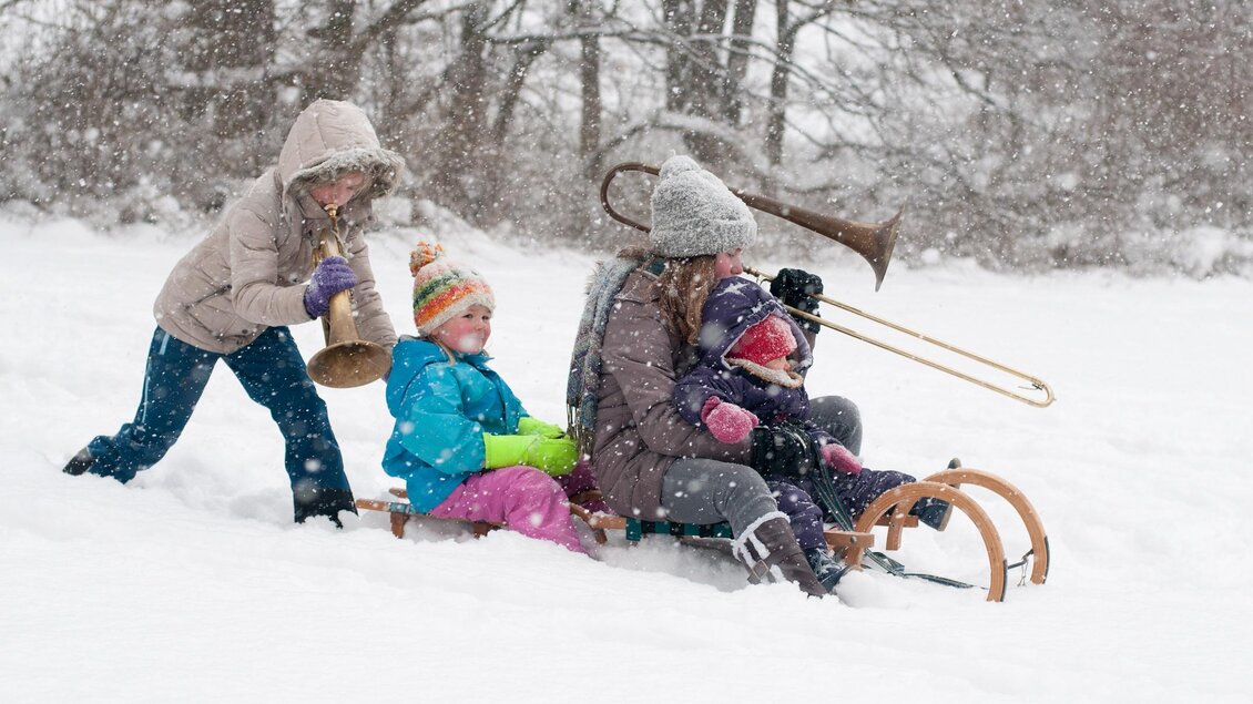 Eine Gruppe von Kindern und Erwachsenen spielt im Schnee. Einige fahren Schlitten, während andere Musikinstrumente spielen. | © (c)STVLW_Unterrainer