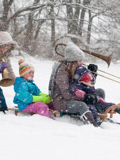 A group of children and adults is playing in the snow. Some are sledding while others are playing musical instruments. | © (c)STVLW_Unterrainer