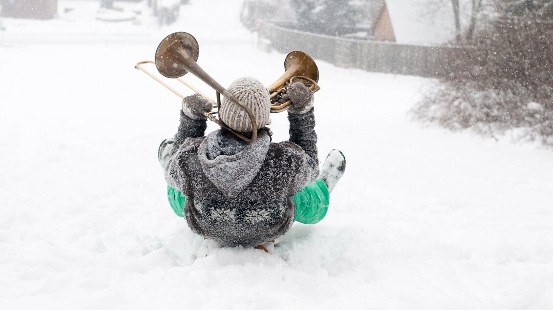 Ein Mensch sitzt im Schnee und hält eine Posaune. Es schneit stark und die Umgebung ist winterlich. | © Steirisches Volksliedwerk - Unterrainer