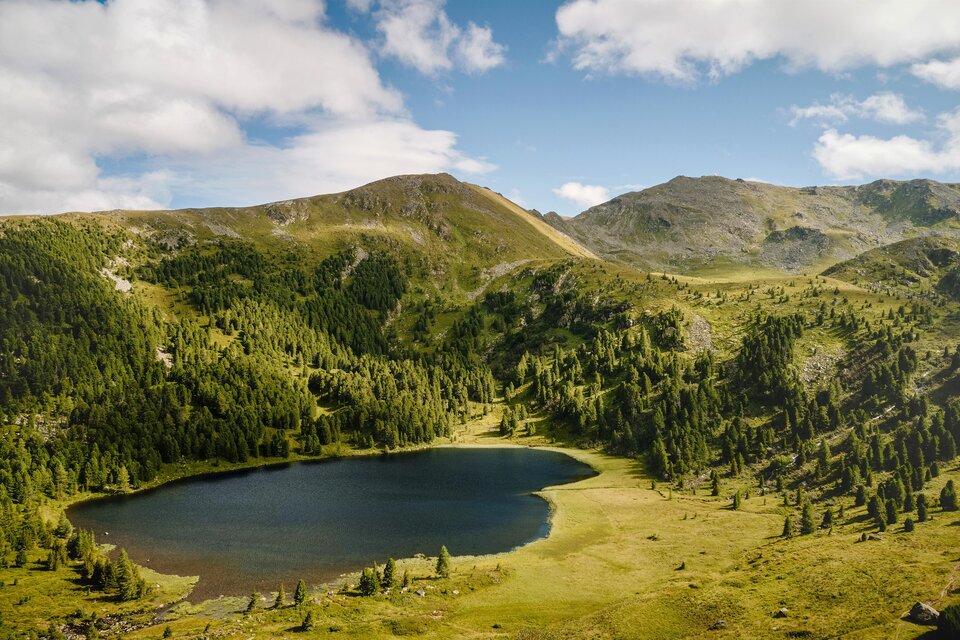 Ein malerischer See umgeben von grünen Wäldern und sanften Hügeln. Der Himmel ist klar mit wenigen Wolken. | © Michael Königshofer