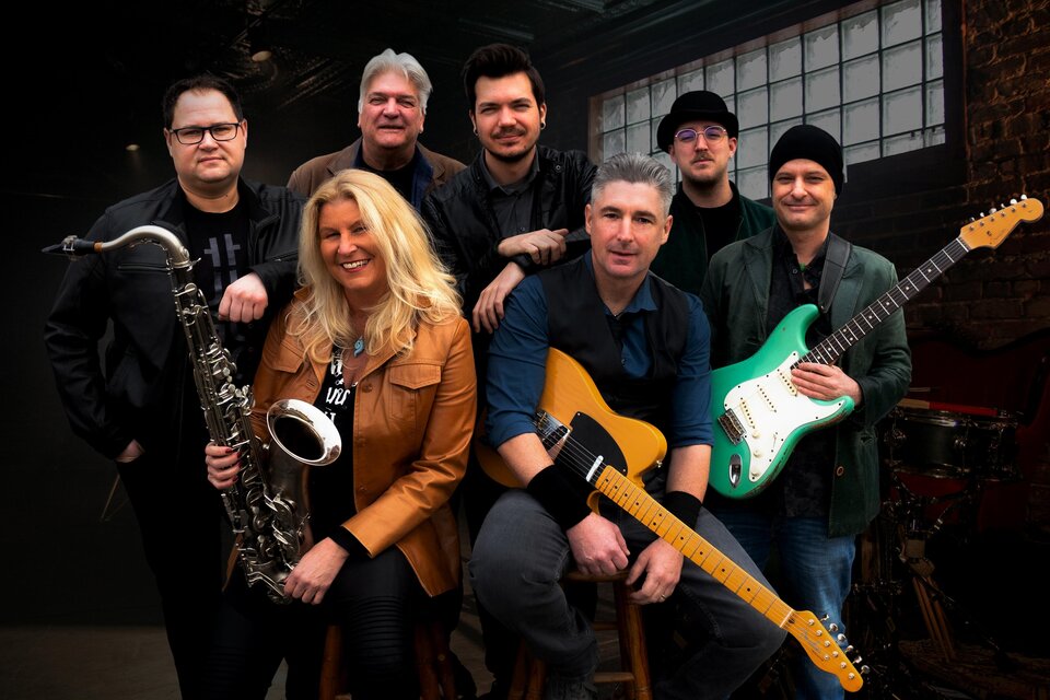 Eine Musikergruppe bestehend aus sieben Personen, die verschiedene Instrumente halten. Sie lächeln in einem gemütlichen Raum mit einem großen Fenster. | © BOSStria