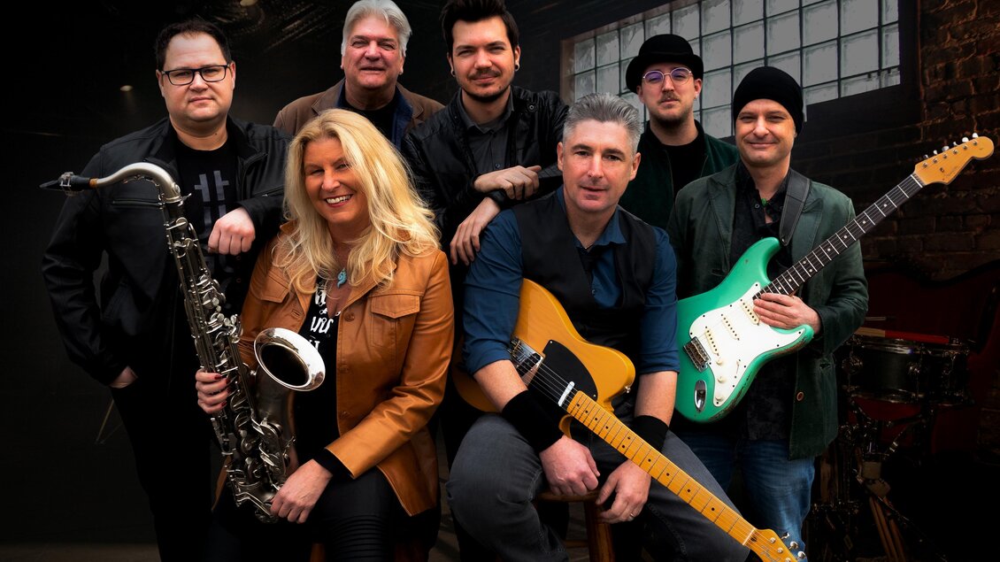 Eine Musikergruppe bestehend aus sieben Personen, die verschiedene Instrumente halten. Sie lächeln in einem gemütlichen Raum mit einem großen Fenster. | © BOSStria