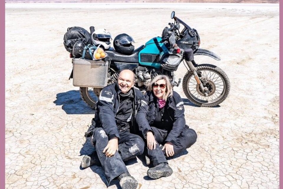 Ein Paar sitzt lächelnd auf dem trockenen Boden neben einem Motorrad. Im Hintergrund sind trockene Landschaft und sanfte Hügel zu sehen. | © Reinhold Ambros