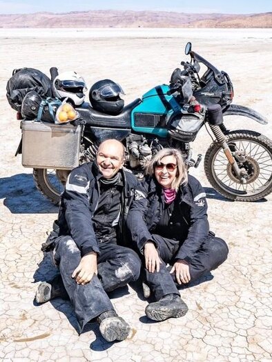 A couple sits smiling on the dry ground next to a motorcycle. In the background, dry landscape and gentle hills can be seen. | © Reinhold Ambros