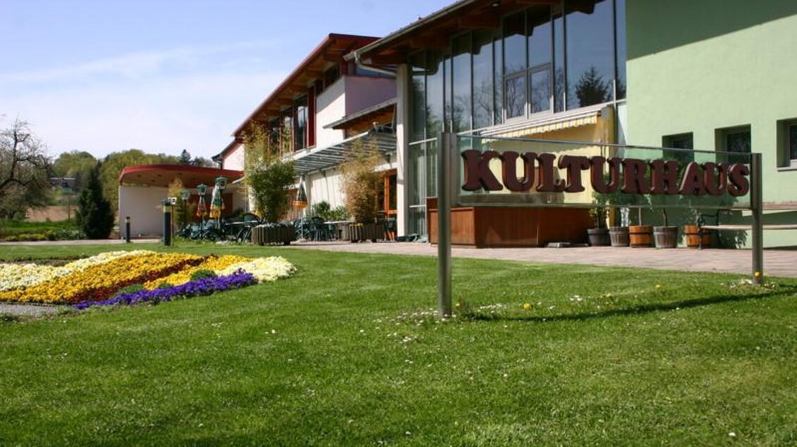 Ein modernes Kulturhaus mit großen Fenstern und einem einladenden Eingangsbereich. Im Vordergrund sind schön angelegte Blumenbeete zu sehen. | © Lederer