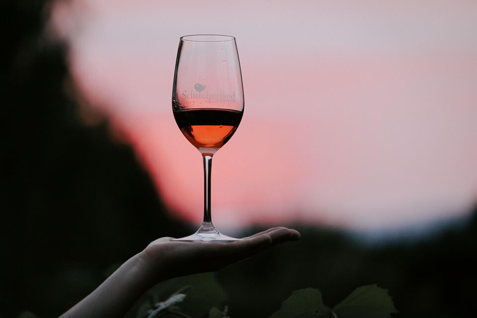 Ein Glas Roséwein wird von einer Hand gehalten. Im Hintergrund ist ein sanfter Sonnenuntergang zu sehen. | © Schilcherland Steiermark