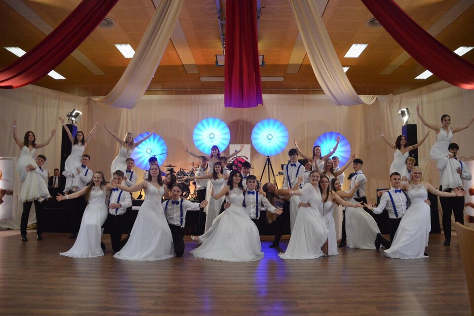 Eine Gruppe von jungen Menschen in festlicher Kleidung feiert auf einer Bühne. Im Hintergrund sind farbenfrohe Lichter und Dekorationen zu sehen. | © Stadtkapelle Fehring