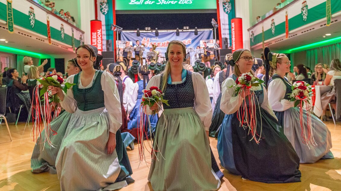 Eine festliche Veranstaltung mit mehreren Frauen, die traditionelle Trachtenkleider tragen. Im Hintergrund sieht man eine Bühne mit einer Band und viele Zuschauer. | © Picwish, Robert Tüchi