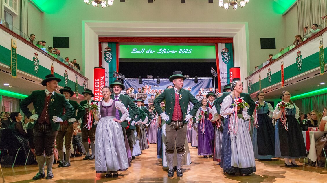 Tänzer in traditioneller Tracht führen einen Volkstanz auf. Der Saal ist festlich dekoriert mit grünen und roten Farben. | © Picwish, Robert Tüchi