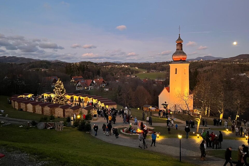 Menschen besuchen den Bartholomäer Adventmarkt mit Kirche und Lichtern in der Dämmerung. | © Josef Birnstingl