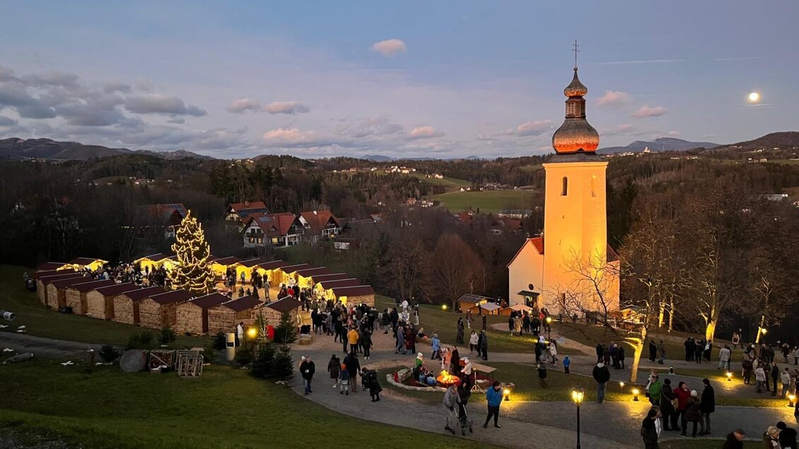 Menschen besuchen den Bartholomäer Adventmarkt mit Kirche und Lichtern in der Dämmerung. | © Josef Birnstingl
