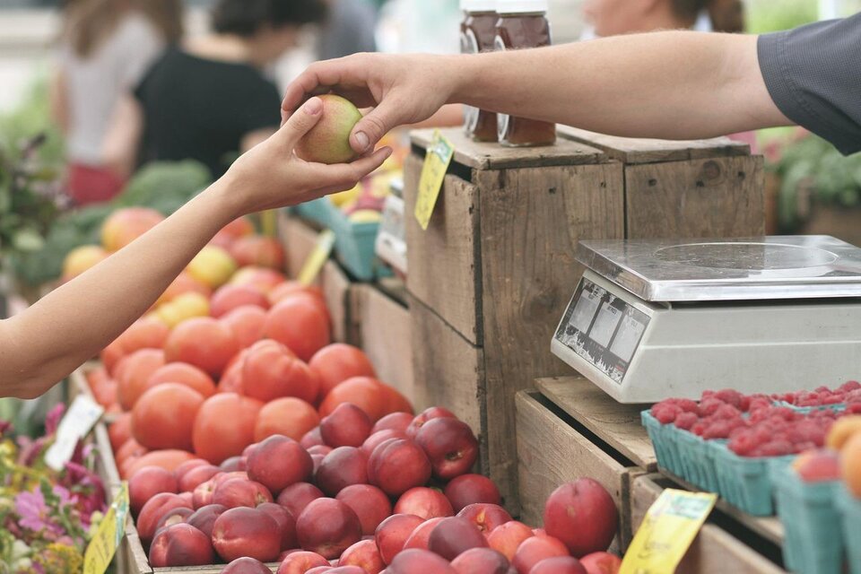 Ein Marktstand mit frischem Obst, darunter Äpfel und Pfirsiche. Eine Hand reicht einen Apfel an einen Verkäufer. | © google Beispielfoto