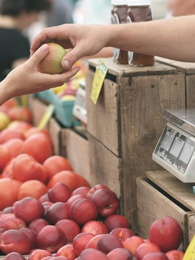 Ein Marktstand mit frischem Obst, darunter Äpfel und Pfirsiche. Eine Hand reicht einen Apfel an einen Verkäufer. | © google Beispielfoto