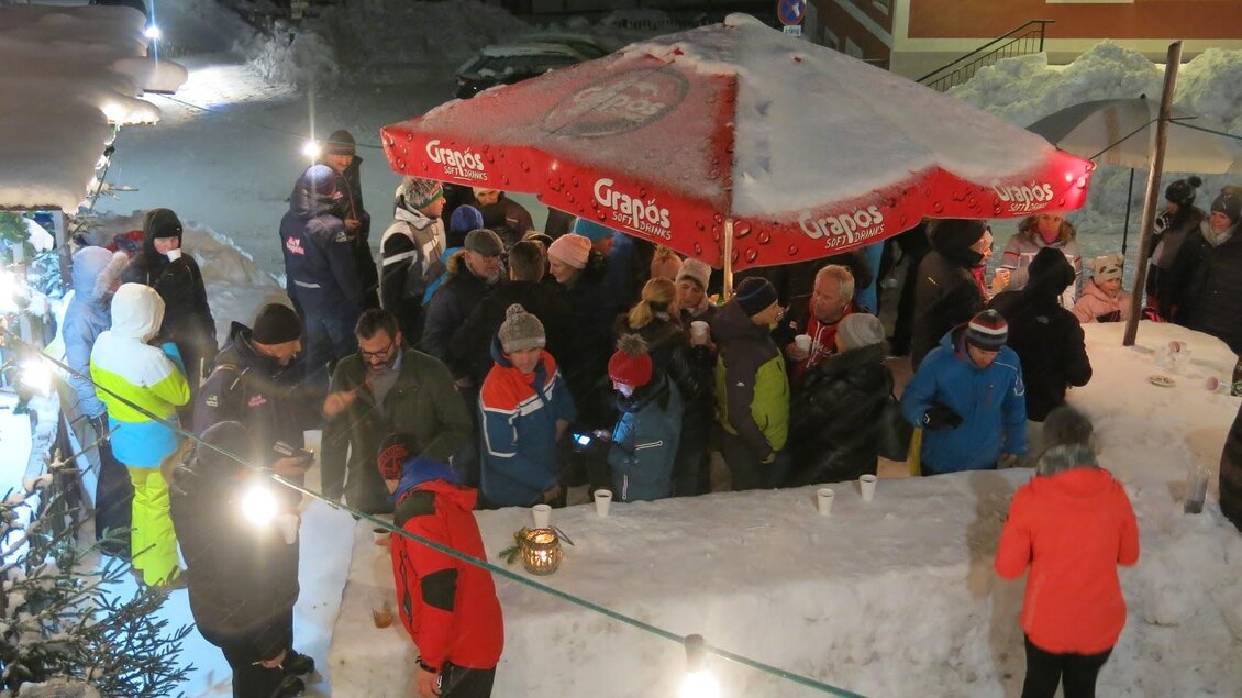 Eine belebte winterliche Szene mit vielen Menschen, die sich unter einem roten Schirm versammeln. Der Boden ist mit Schnee bedeckt und es gibt festliche Lichter. | © Bergrettung