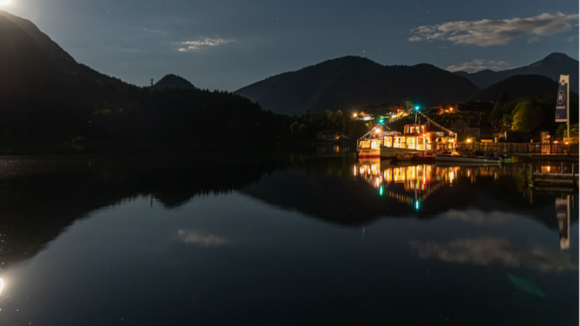 Ein ruhiger See bei Nacht, umgeben von Bergen. Das Licht reflektiert sich auf der Wasseroberfläche und schafft eine malerische Stimmung. | © Schifffahrt Grundlsee