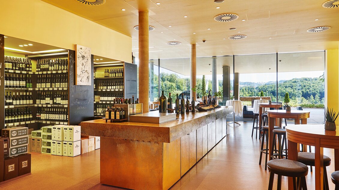Eine moderne Weinbar mit einer eleganten Holztheke und großen Fenstern, die einen Blick auf die umliegende Landschaft bieten. Regal mit Weinflaschen ist im Hintergrund sichtbar. | © LOISIUM Südsteiermark
