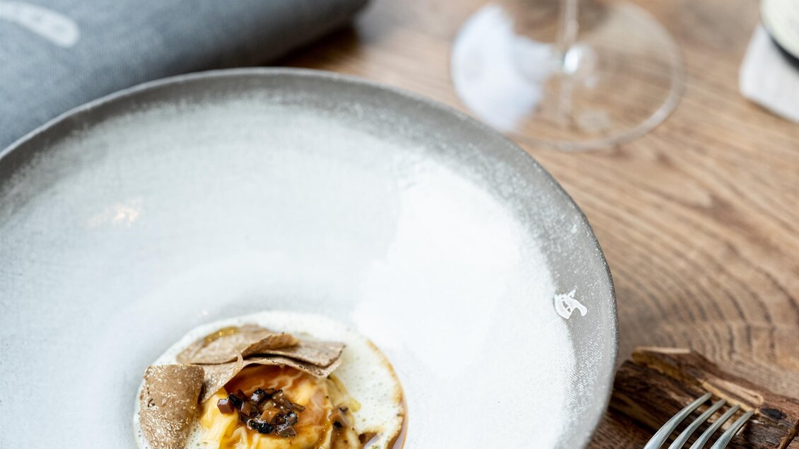 Ein stilvolles Gericht in einer eleganten Schüssel auf einem Holztisch. Im Hintergrund steht ein Glas Wein und ein graues Servietten. | © Michael Reidinger