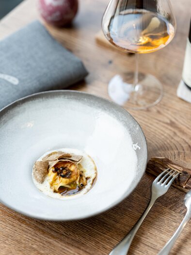 A stylish dish in an elegant bowl on a wooden table. In the background, there is a glass of wine and a gray napkin. | © Michael Reidinger