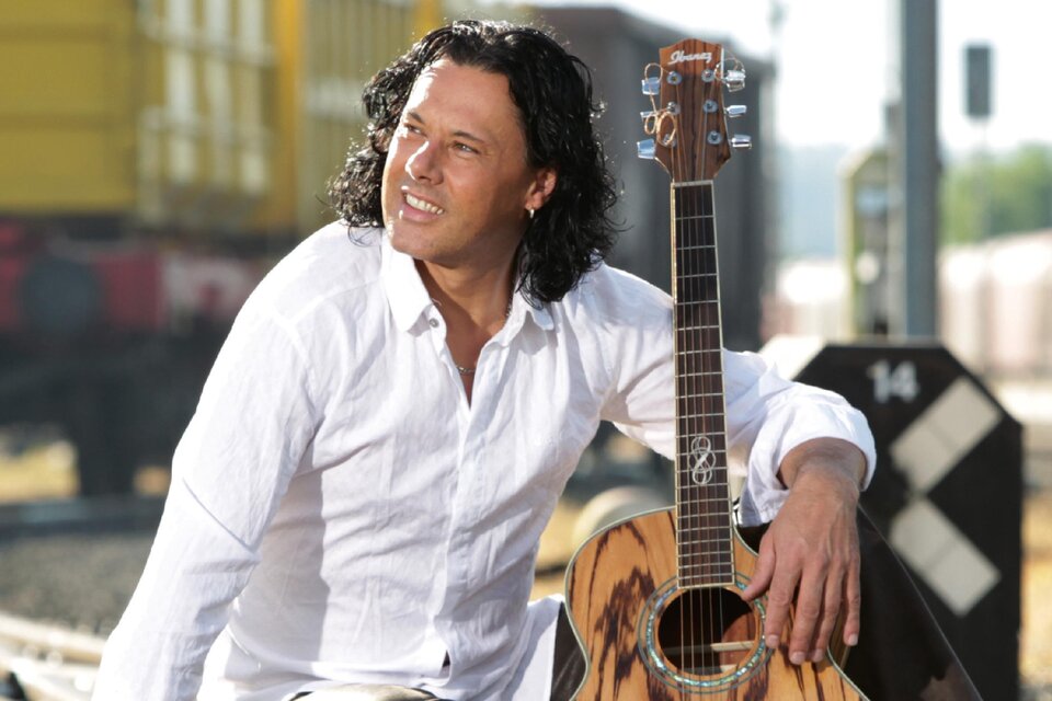 Ein Mann mit lockigem Haar sitzt an einem Bahngleis und hält eine Akustikgitarre. Er trägt ein weißes Hemd und schaut lächelnd in die Ferne. | © Vitalhotel der Parktherme