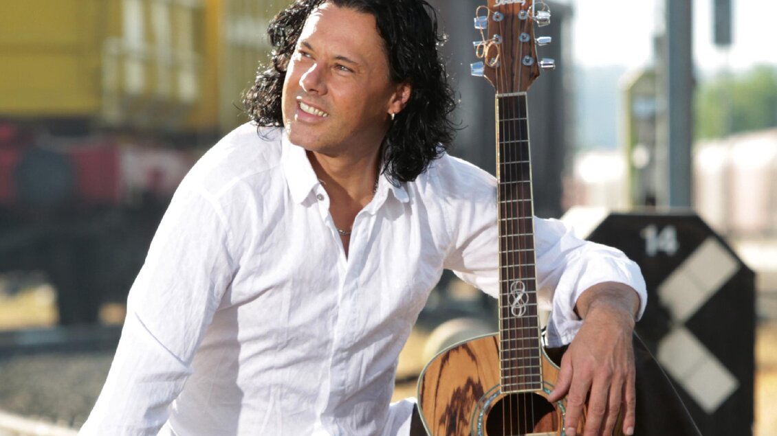 Ein Mann mit lockigem Haar sitzt an einem Bahngleis und hält eine Akustikgitarre. Er trägt ein weißes Hemd und schaut lächelnd in die Ferne. | © Vitalhotel der Parktherme