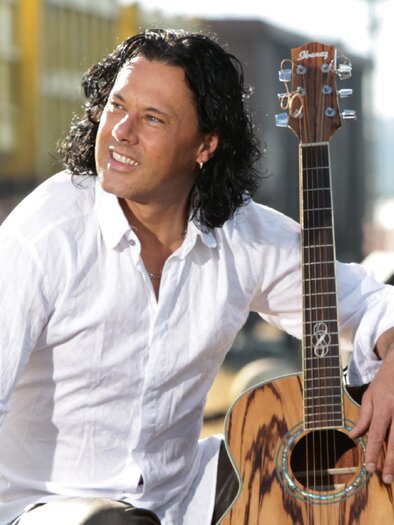 Ein Mann mit lockigem Haar sitzt an einem Bahngleis und hält eine Akustikgitarre. Er trägt ein weißes Hemd und schaut lächelnd in die Ferne. | © Vitalhotel der Parktherme