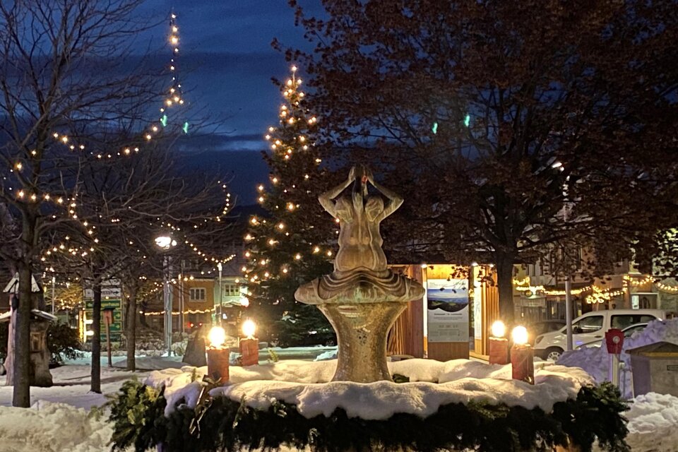 Ein schöner Brunnen im Winter, umgeben von frisch gefallenem Schnee. Lichter schmücken die Bäume und schaffen eine festliche Atmosphäre.