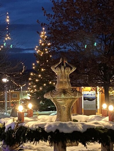 Ein schöner Brunnen im Winter, umgeben von frisch gefallenem Schnee. Lichter schmücken die Bäume und schaffen eine festliche Atmosphäre.