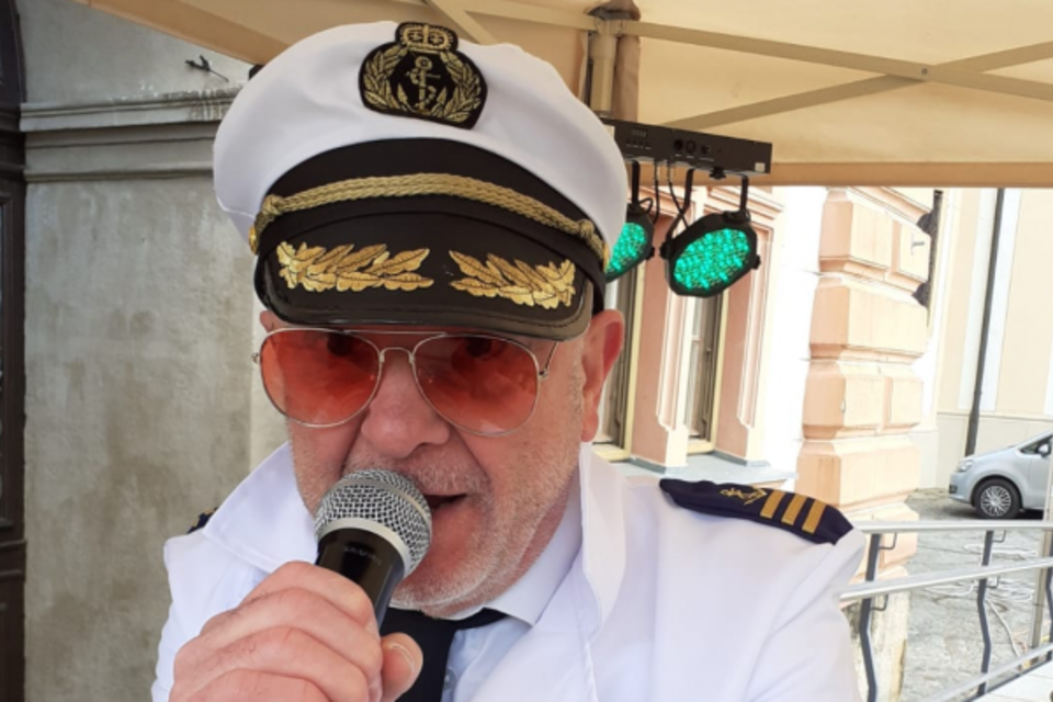 Ein Mann im Kapitänsoutfit mit einer Sonnenbrille hält ein Mikrofon. Er spricht anscheinend zu einem Publikum und steht unter einem Zelt.