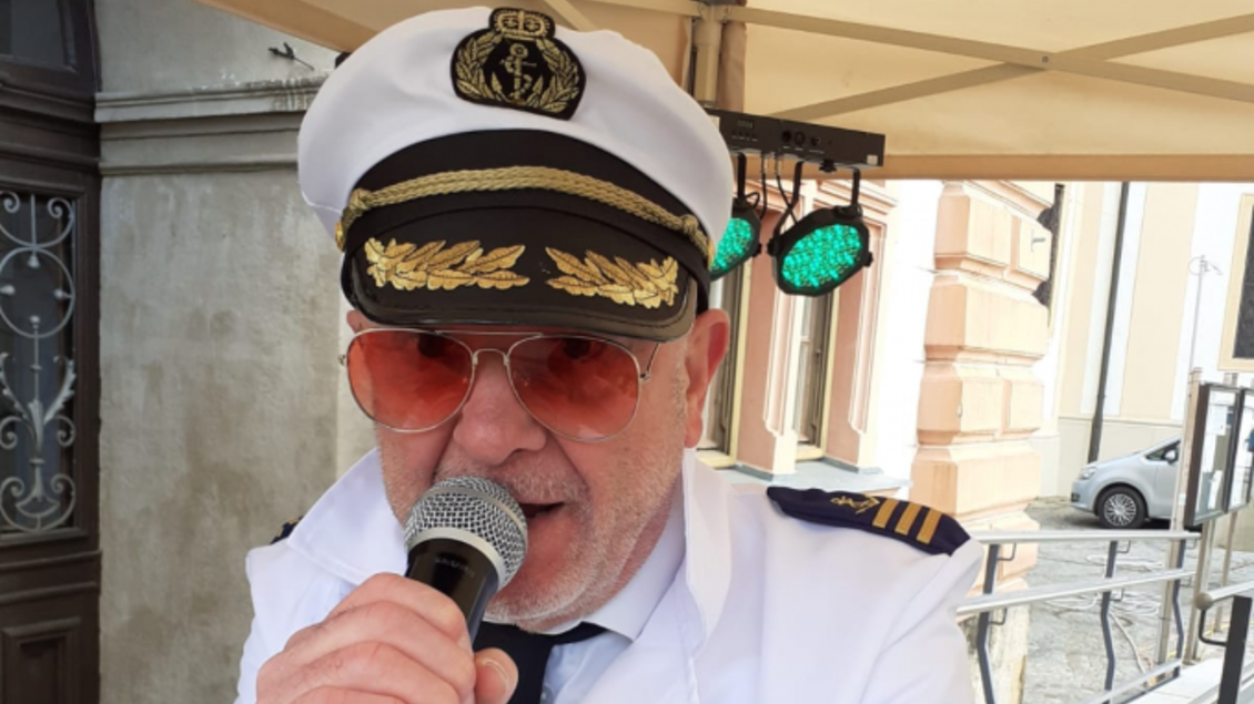 Ein Mann im Kapitänsoutfit mit einer Sonnenbrille hält ein Mikrofon. Er spricht anscheinend zu einem Publikum und steht unter einem Zelt.