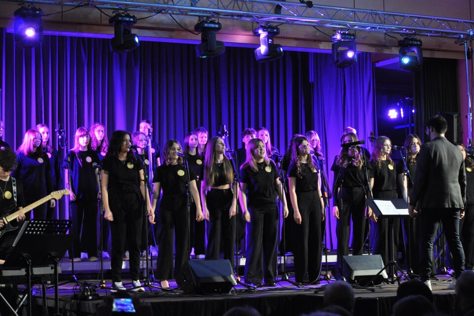 Ein Chor aus jungen Sängerinnen und Sängern in schwarzer Kleidung steht auf einer Bühne. Bunte Lichter und Mikrofone schaffen eine festliche Atmosphäre. | © BORG Bad Radkersburg