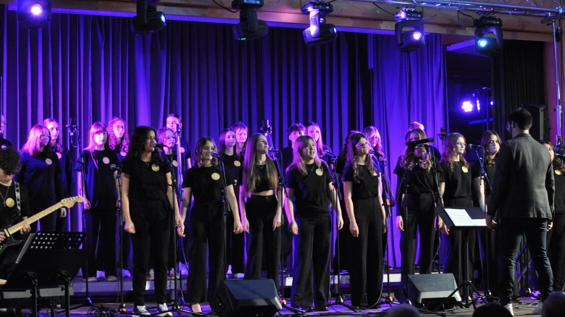 Ein Chor aus jungen Sängerinnen und Sängern in schwarzer Kleidung steht auf einer Bühne. Bunte Lichter und Mikrofone schaffen eine festliche Atmosphäre. | © BORG Bad Radkersburg