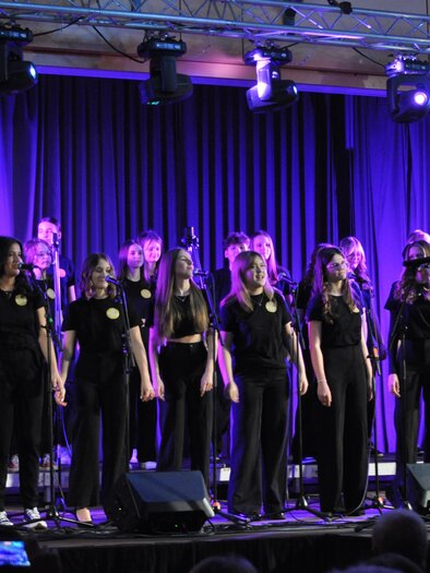 A choir of young singers in black clothing stands on a stage. Colorful lights and microphones create a festive atmosphere. | © BORG Bad Radkersburg