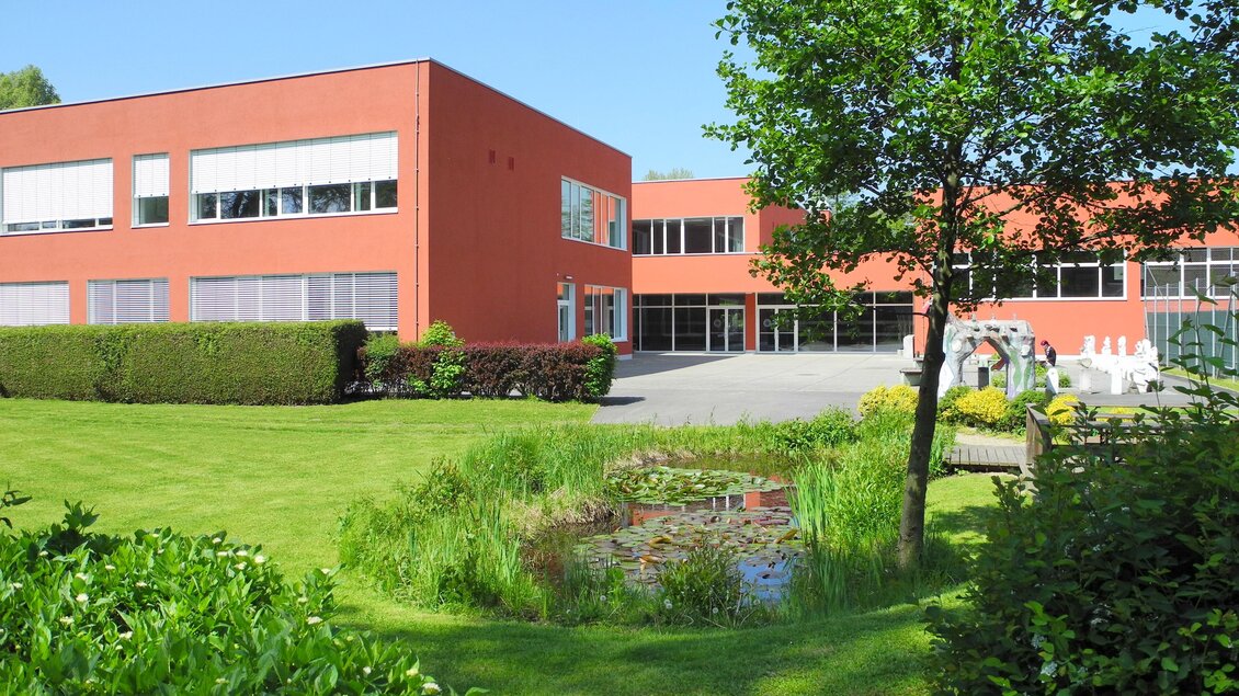 Ein modernes Gebäude mit roten Wänden, umgeben von gepflegten Grünflächen. Im Vordergrund befindet sich ein kleiner Teich und Bäume. | © BORG Bad Radkersburg