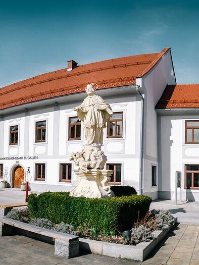 Ein historisches Gebäude mit rotem Dach und großen Fenstern. Vor dem Gebäude steht eine Statue auf einem Podest. | © Stefan Leitner