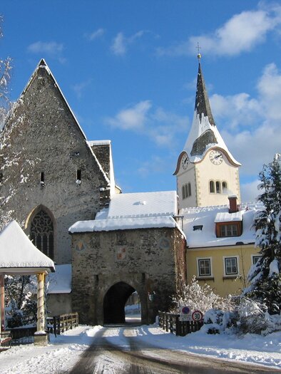 Ein malerisches Winterdorf mit schneebedeckten Dächern und einer Kirche mit Turm. Die Landschaft wirkt ruhig und einladend.