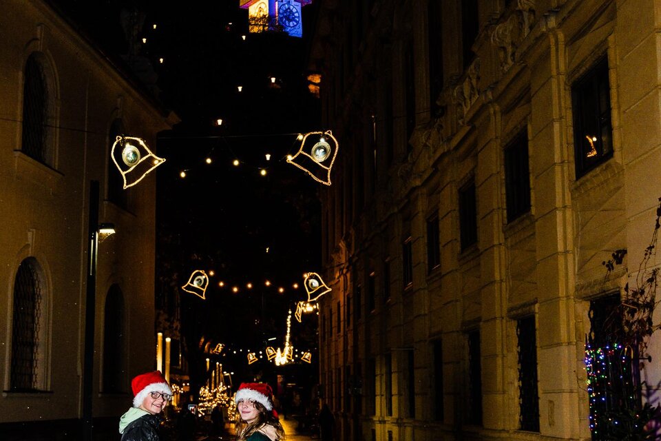 Zwei Personen mit Weihnachtsmützen stehen in einer festlich beleuchteten Straße. Der Weg ist nass und glänzt, während Lichter an den Gebäuden funkeln. | © (c) Region Graz - Mias Photoart