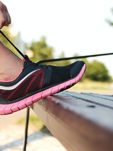A foot in a black and pink sports shoe is tying a shoelace. In the background, a park-like environment with trees is visible. | © Bild: Pexels