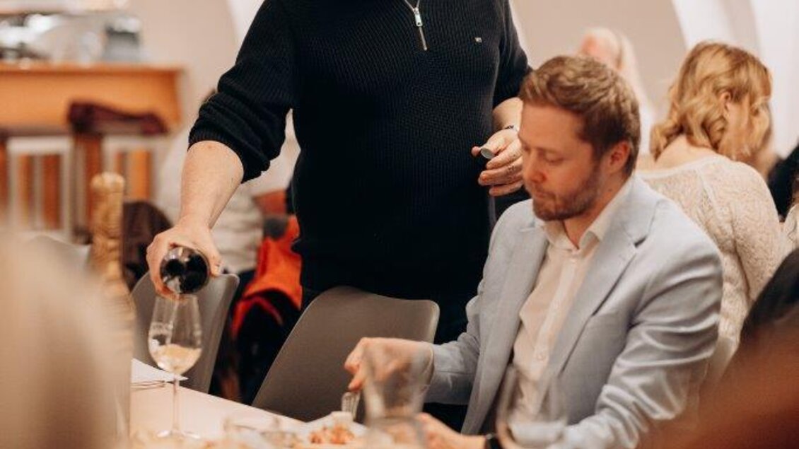 Ein Mann serviert Wein an einen Tisch in einem Restaurant. Im Hintergrund sind andere Gäste und eine gemütliche Atmosphäre zu sehen. | © Christoph Draxl