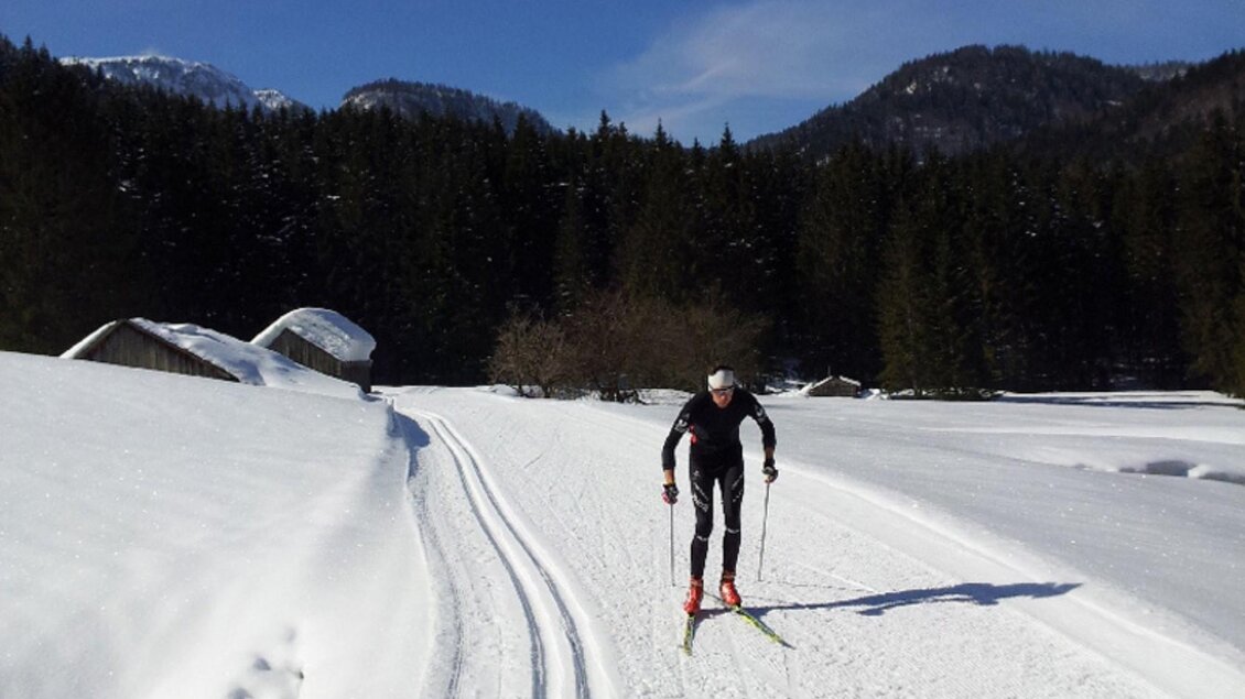 Ein Skilangläufer fährt durch eine schneebedeckte Landschaft. Im Hintergrund sind Berge und Nadelbäume sichtbar. | © Georg Petritsch