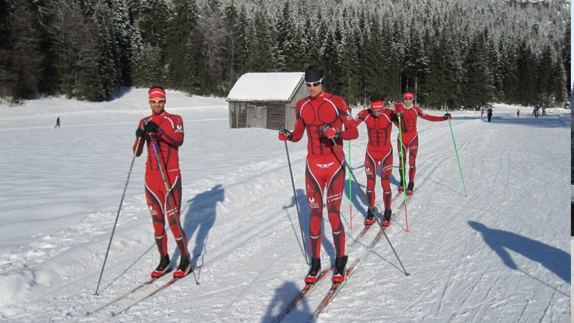 Eine Gruppe von Skilangläufern in roten Anzügen fährt auf einer verschneiten Strecke. Im Hintergrund sind Bäume und ein kleiner Holzschuppen zu sehen. | © Georg Petritsch