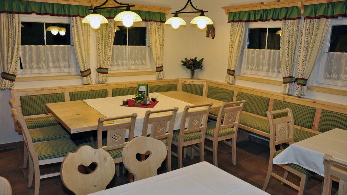 Ein gemütlicher Essbereich mit einem großen Tisch und Bänken. Die Wände sind mit Vorhängen und dekorativen Elementen in traditionellem Stil gestaltet. | © Poppnangerl