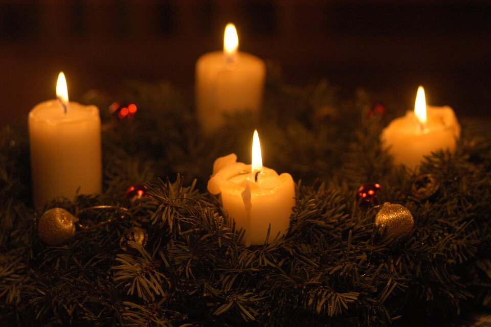 Ein Adventskranz mit vier brennenden, weißen Kerzen. Er ist mit Tannenzweigen und kleinen dekorativen Elementen geschmückt.