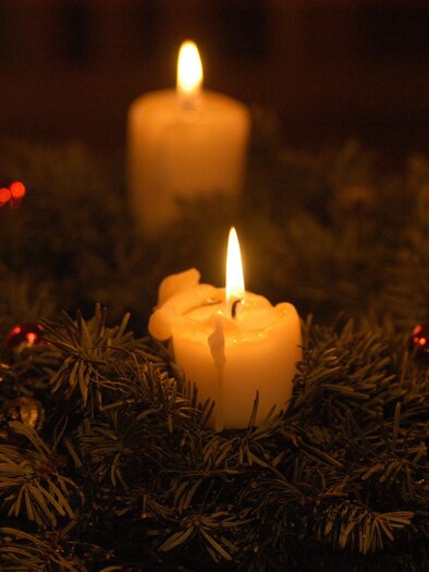 An Advent wreath with four burning white candles. It is decorated with fir branches and small decorative elements.