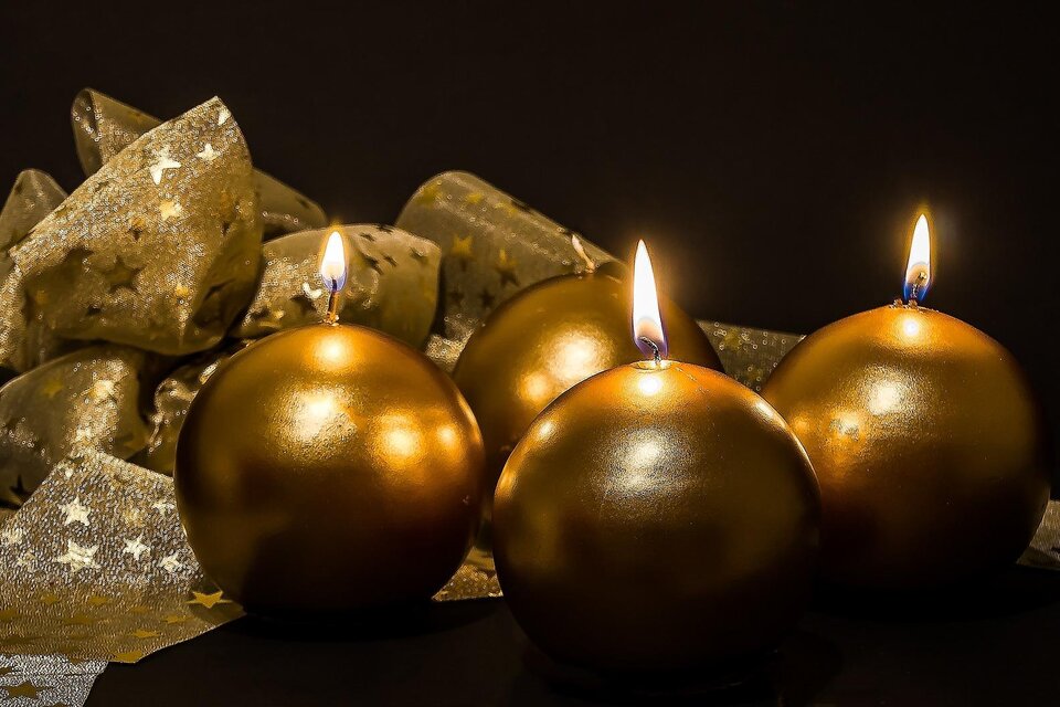 Vier goldene Kerzen brennen auf einem dunklen Hintergrund. Daneben liegt eine goldene Schleife mit Sternen. | © Pixabay